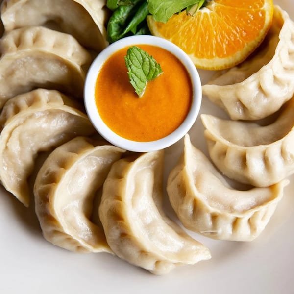 Nepalese momos arranged on a white plate with a side of orange dipping sauce and an orange slice.